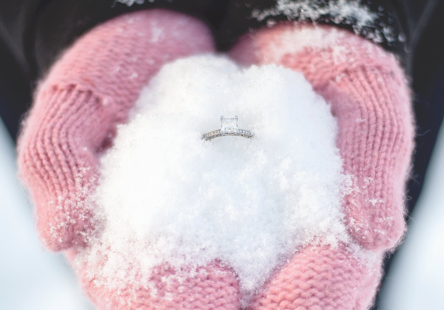An engagement ring in snow held by pink knitted gloves